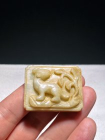 明 和田玉籽料 白玉沁色 瑞兽纹 带饰 把件 挂件 宽4.2厘米 高3.3厘米 厚1.2厘米 重28.4克 和田籽料为材，微微带黄沁渍，古朴典雅，甚具玩味。长方形带扣，高浮雕瑞兽纹，兽呈蹲卧回首状，兽耳伏贴侧边，独角后翘，四肢粗壮有力、屈曲收于身下，一旁饰山石草木图案，本器两侧有一长方形孔对穿，可供穿系用。