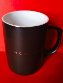 【LINCOLN林肯陶瓷马克杯】进口林肯车原车配送，有柄，黑釉。高11.0厘米，通长（含柄）11.0厘米，直径8.0厘米，厚度0.3厘米，质量390.0克。杯口金线饰边。杯体印林肯车标及英文“LINCOLN”。全新，尚未使用，带原包装盒。