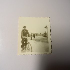 老照片–青年在乡村路上骑自行车留影（身后有多名村民）