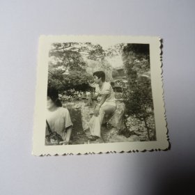 老照片–帅气青年坐在景区大石头上留影（左侧有一名女游客，身后亭台树木清晰可见）